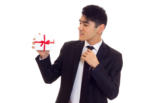 Eleganter Mann mit schwarzen Haaren in weißem Hemd und schwarzem Anzug mit Krawatte mit einem Geschenk auf weißem Hintergrund