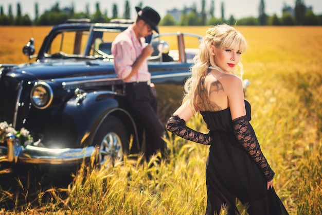 Eleganter Mann und Frau, die auf einem Weizenfeld in der Nähe eines altmodischen Autos stehen