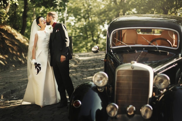 Eleganter, stilvoller Bräutigam, der sanft wunderschöne Braut im schwarzen Auto im Licht im Park küsst, ungewöhnliches Luxus-Hochzeitspaar im romantischen Moment im Retro-Stil