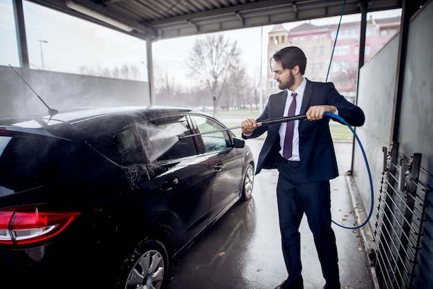 Eleganter stilvoller junger fokussierter Mann in einer Anzugreinigungsseite des Autos mit einer Wasserpistole auf Selbstbedienungswaschautostation.