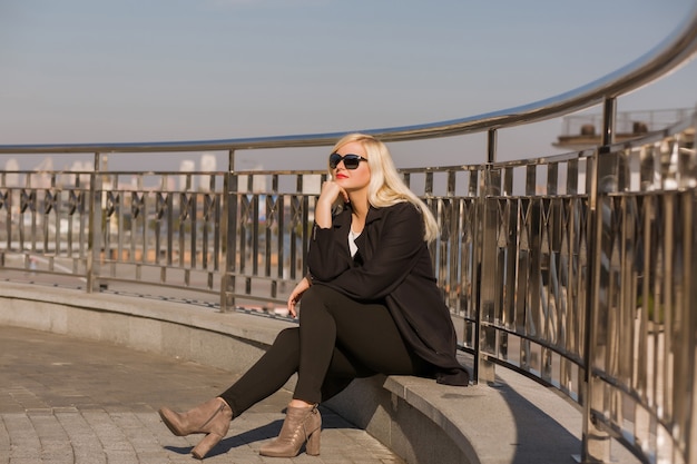 Elegantes blondes Model, das im schwarzen Mantel posiert, trägt eine Sonnenbrille auf dem Hintergrund der Stadt