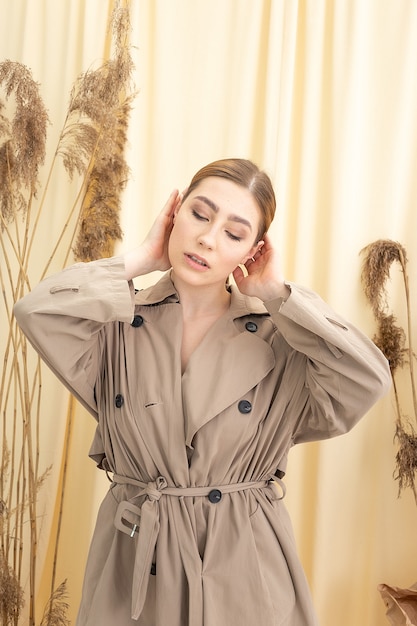 Elegantes kaukasisches hübsches Mädchen im beige Regenmantel mit dem langen Haar, das auf Pastellstoff mit getrockneten Blumen aufwirft