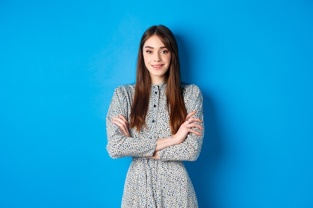 Elegantes kaukasisches Mädchen im Kleid verschränkt die Arme auf der Brust und steht auf Blau.