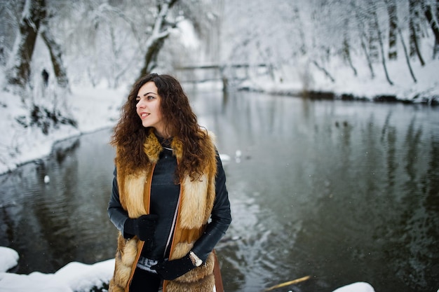 Elegantes lockiges Mädchen im Pelzmantel im verschneiten Waldpark gegen gefrorenen Fluss im Winter.