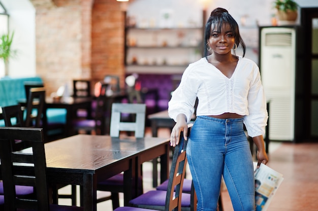 Elegantes mujeres afroamericanas en blusa blanca y pantalones vaqueros azul posando en el café con periódico.