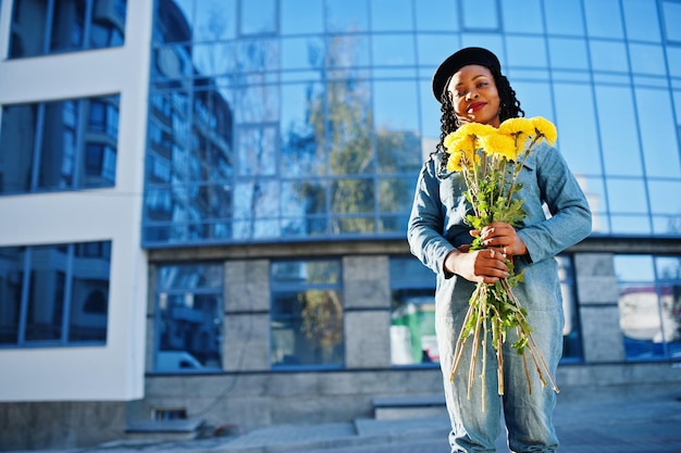 Elegantes mujeres afroamericanas de moda en jeans y boina negra con ramo de flores amarillas posadas al aire libre en un día soleado contra un edificio moderno azul