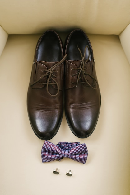 Elegantes zapatos clásicos para hombre con cordones, pajarita a cuadros y gemelos sobre fondo beige. Accesorios para el novio en la boda.