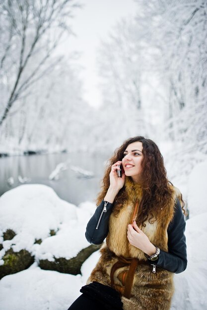 Eleganz lockiges Mädchen im Pelzmantel mit Handy im verschneiten Waldpark im Winter