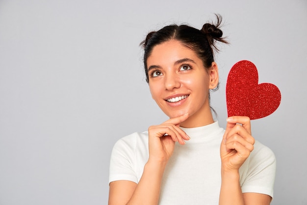 Elegir la variante adecuada. Retrato de mujer sosteniendo un corazón rojo brillante Valentine pensando mientras sonríe