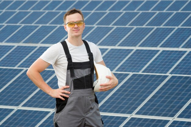 Elektrischer Arbeiter, der weißen Sicherheitshut hält und am Kraftwerk steht. Solaringenieur in gelben Schutzgläsern und grauen Overalls, die in der Nähe des Sonnenkollektorfeldes stehen.