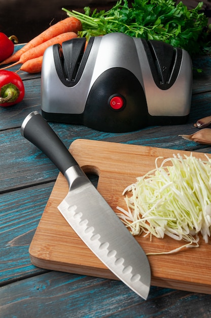 Elektrischer Messerschärfer. Der Kunststoffkörper ist grau-schwarz. Im Vordergrund steht ein Messer und gehackter Kohl auf einem Holzbrett. Im Hintergrund Gemüse und Kräuter.