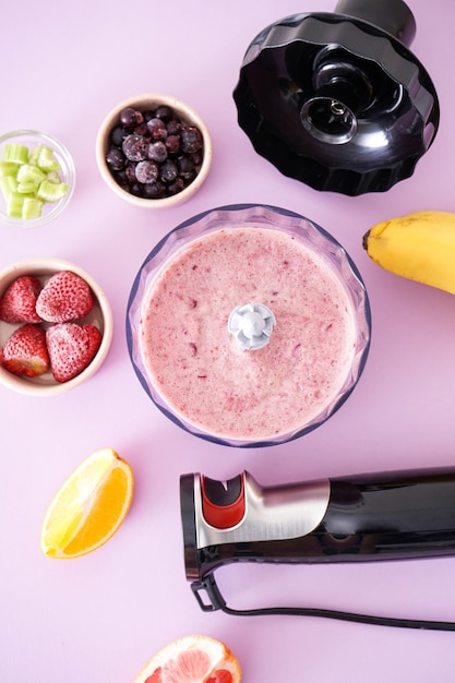 Foto elektrischer stabmixer und zubehör aus schwarzem kunststoff mit früchten und smoothie auf violettem hintergrund, vertikal, flach gelegt.