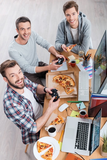 Eles gostam do que fazem. Vista superior de três jovens jogando no computador e comendo pizza enquanto estão sentados à mesa