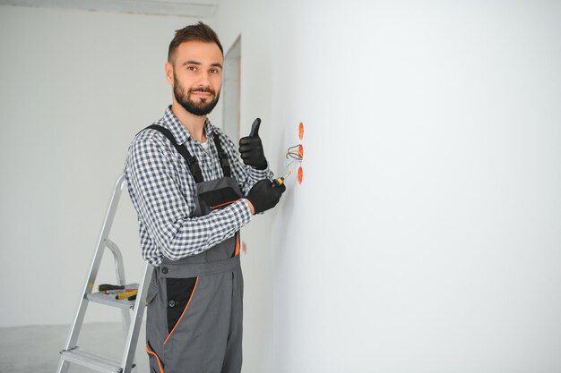 Eletricista de uniforme montando tomadas elétricas na parede branca dentro de casa