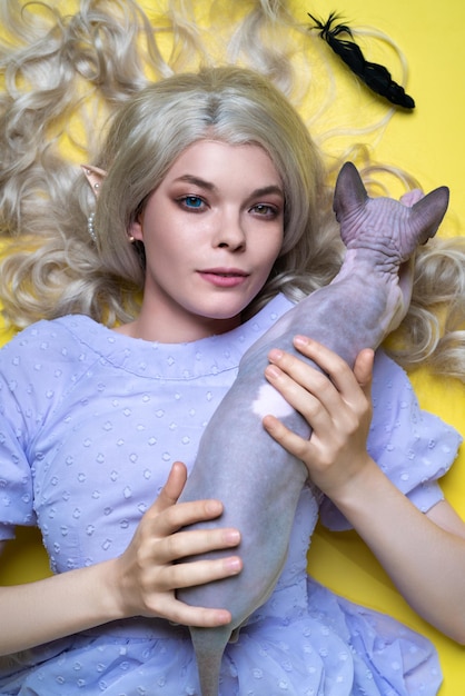 Elfo de cosplay de mujer joven en vestido azul mirando a la cámara acostada con gato sobre fondo amarillo