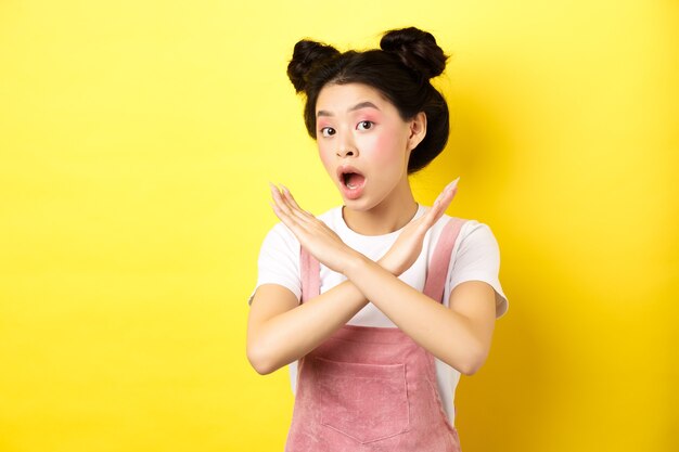 Ella dice que no. Chica de belleza asiática con maquillaje, mostrando signo de cruz, parada y oferta de rechazo, de pie decepcionada en amarillo.