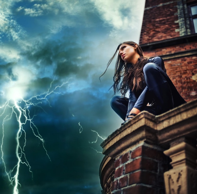 Foto ella es una fuerza de la naturaleza superhéroe mujer joven seductora en un catsuit negro agazapada en una azotea en una tormenta eléctrica