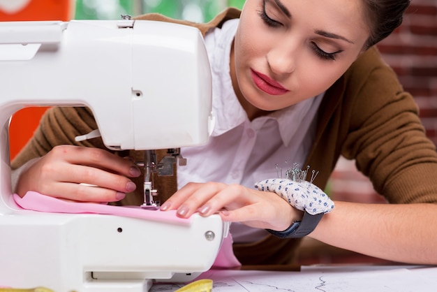 Ella es una sastre experta. Diseñadora de moda femenina confiada que trabaja en la máquina de coser mientras está sentada en su lugar de trabajo