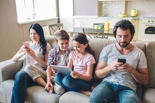 Eltern sitzen mit Kindern auf dem Sofa und schauen auf Telefone. Kinder sind zwischen Frau und Mann. Mädchen hält Tablette in ihren Händen. Sie schauen auf den Bildschirm.