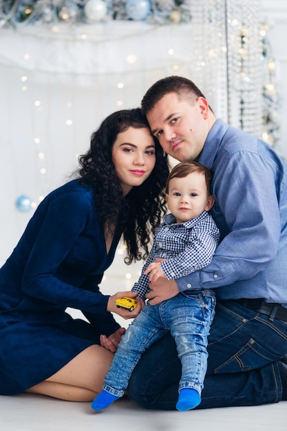 Eltern und kleiner Sohn sitzen auf dem Boden neben dem Kamin und schauen in die Kamera Weihnachtsfotosession