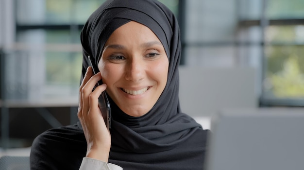 Em close-up, uma jovem mulher árabe sorridente, secretária de hijab, a falar ao telefone no escritório, a consultar um cliente.