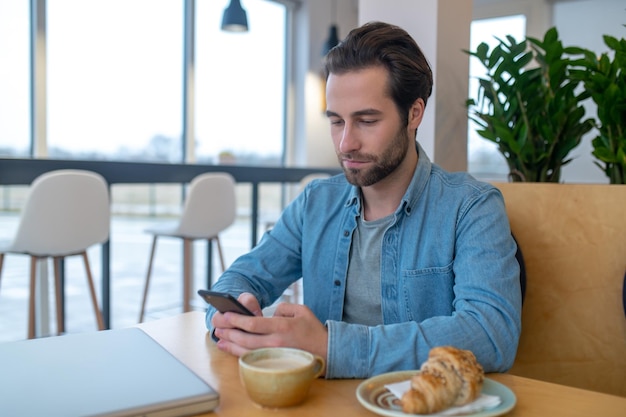 Em formação. Jovem homem caucasiano barbudo olhando para smartphone enquanto está sentado à mesa com o laptop no café durante o dia