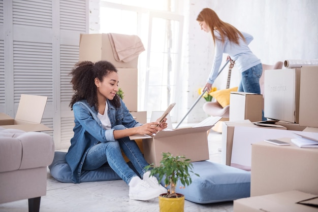 Em novo apartamento. Menina agradável limpando o chão com um esfregão enquanto sua colega de quarto está sentada no chão olhando uma foto emoldurada enquanto desempacota seus pertences