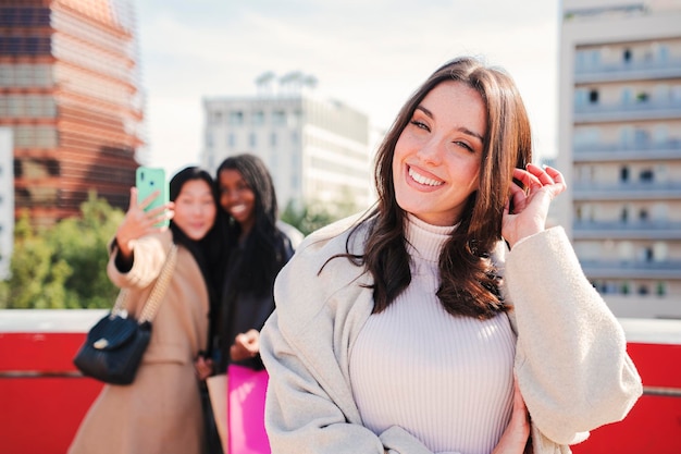 Em primeiro plano, uma jovem e bonita mulher morena com dentes brancos perfeitos sorrindo olhando para a câmera no fundo dois amigos felizes fazendo compras tirando um retrato de selfie