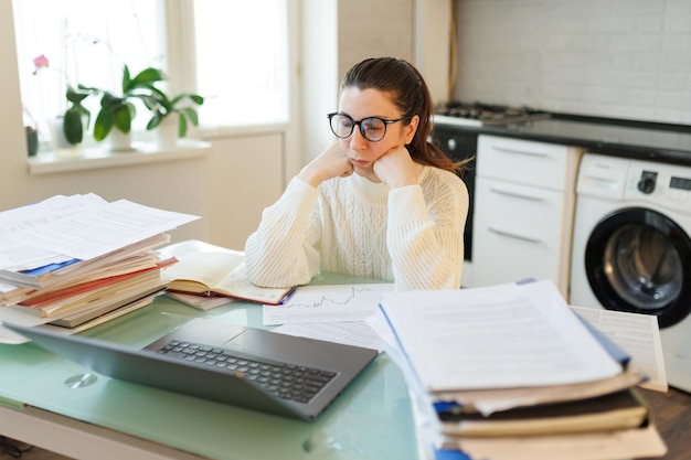 Em seu espaço de trabalho em casa, uma mulher cansada que usa óculos faz uma pausa de seu trabalho em um laptop