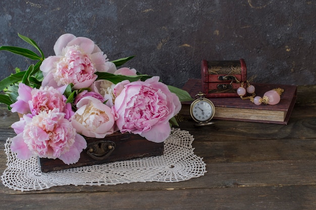 Em um caixão antigo um buquê de peônias rosa, um livro, contas com pedras cor de rosa