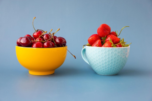 Em um fundo azul há duas tigelas com cerejas e morangos