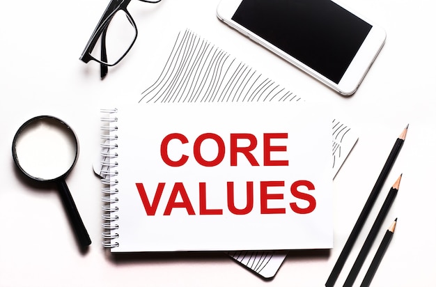 Em um fundo branco óculos, uma lupa, lápis, um smartphone e um caderno com o texto core values