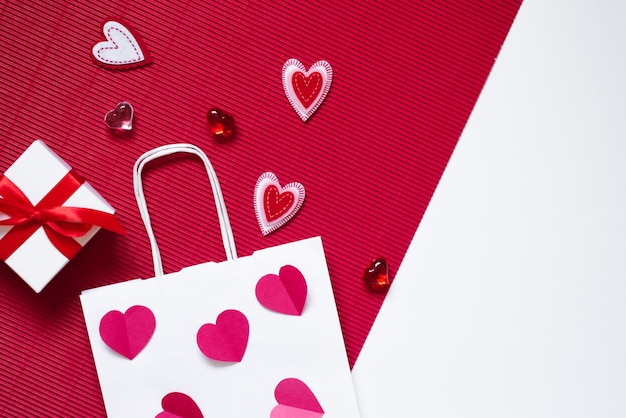 Em um fundo vermelho e branco, uma caixa de presente com um laço rosa, um saco de papel branco, corações de vidro carmesim