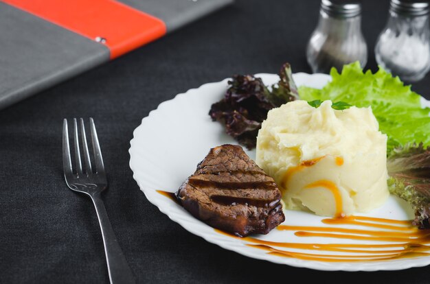 Em um prato branco, em um belo tecido e fundo escuro purê de batata com carne bovina lindamente polida molho balsâmico