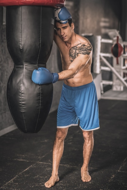 Em uma academia. Homem musculoso em shorts azuis, se exercitando em uma academia e parecendo exausto