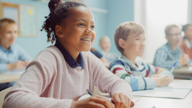 Foto em uma aula de escola primária, retrato de uma brilhante menina negra com aparelhos ortopédicos escreve em um caderno de exercícios, sorri em uma sala de aula júnior com um grupo diversificado de crianças aprendendo coisas novas.