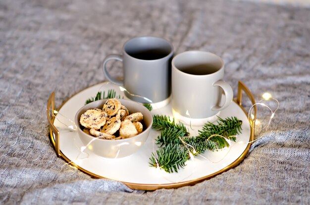 Em uma bandeja de mármore com borda dourada, há duas xícaras de chá cinza e um biscoito de gengibre de chocolate. um deleite para o papai noel. galho de árvore de natal com guirlandas decoram o cartão de ano novo. copie o espaço