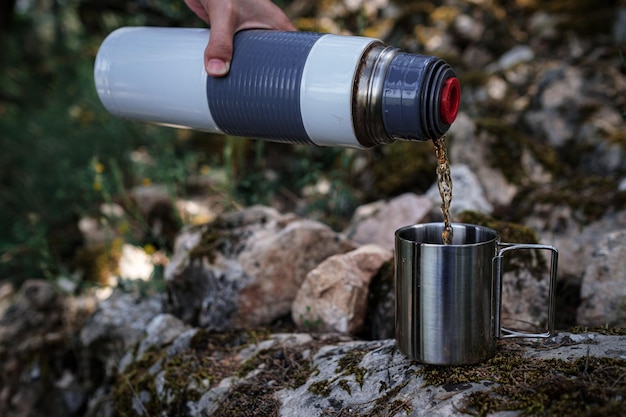 Em uma caneca de acampamento derrama chá de uma garrafa térmica na natureza