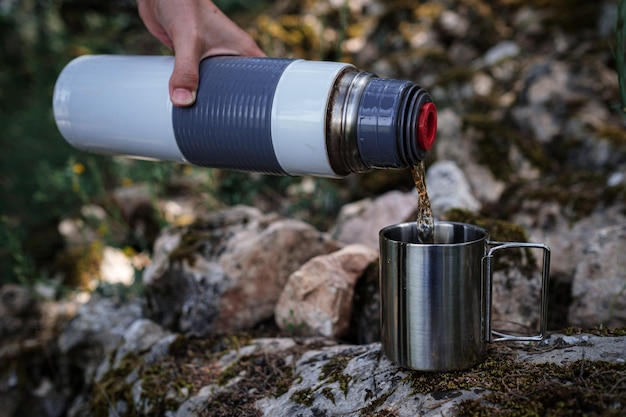 Em uma caneca de acampamento derrama chá de uma garrafa térmica na natureza