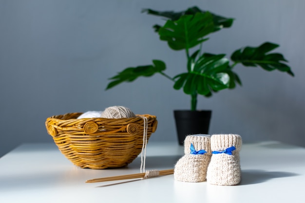 Em uma cesta de vime, há bolas com agulhas de tricô próximas a elas são botinhas de bebê de tricô