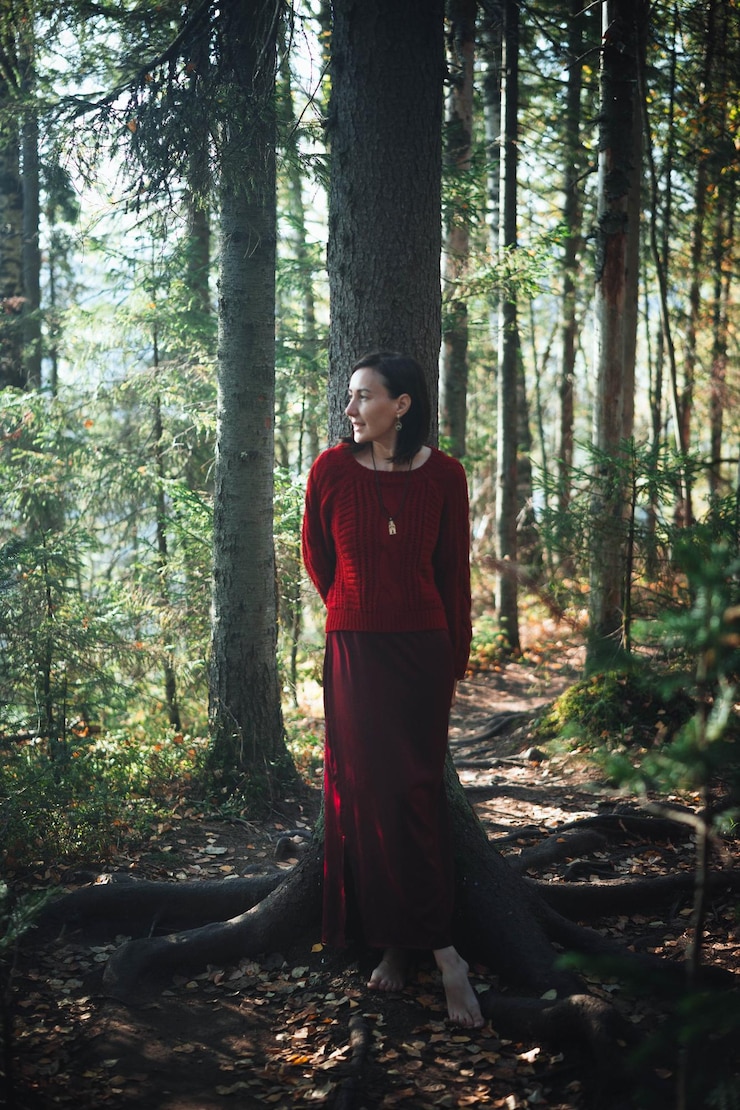 Foto em uma floresta tranquila, uma mulher vestida com um suéter vermelho e uma saia larga se inclina contra uma árvore cercada por vegetação vibrante e luz solar manchada filtrando as folhas.