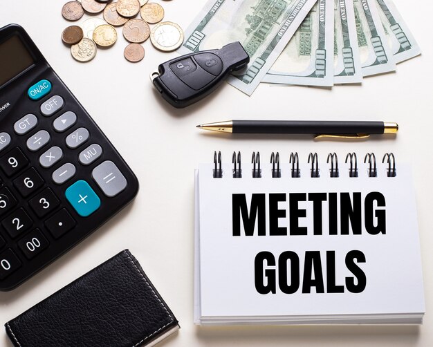 Em uma mesa branca está uma calculadora, a chave do carro, dinheiro, uma caneta e um caderno com a inscrição objetivos da reunião