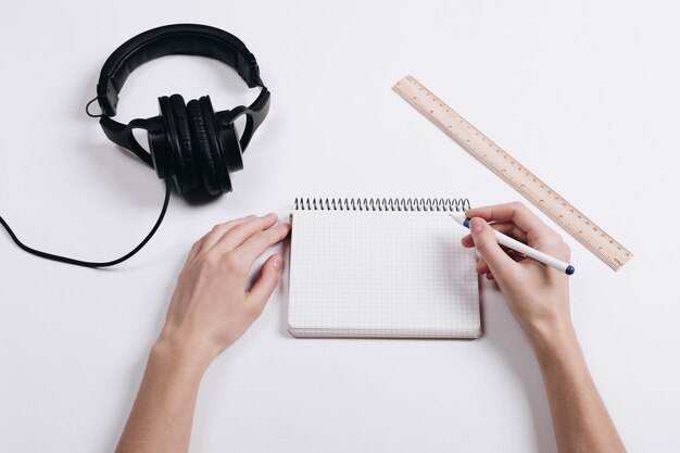 Em uma mesa branca são fones de ouvido, uma régua e um notebook, mãos femininas escrevendo caneta