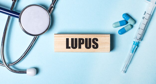 Em uma parede azul, um estetoscópio, uma seringa e comprimidos e um bloco de madeira com a palavra LUPUS. Conceito médico