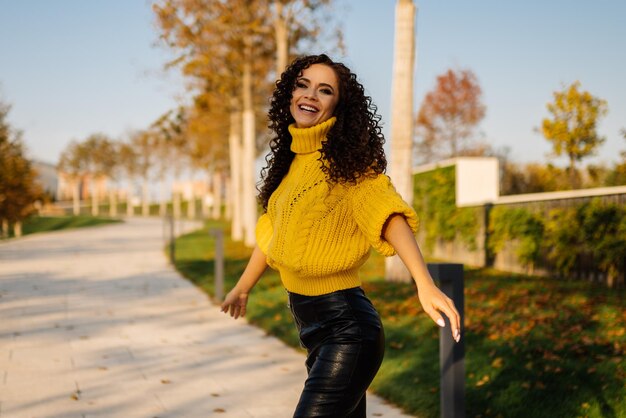 Em uma saia casual e um suéter amarelo quente de tricô grande no caminho do parque vem uma linda mulher e sorri