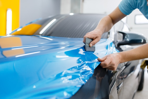 Embalagem de carro, mecânico com rodo instala película protetora de vinil ou filme no capô do veículo. Trabalhador faz detalhamento de automóveis. Revestimento de proteção de pintura automotiva, ajuste profissional