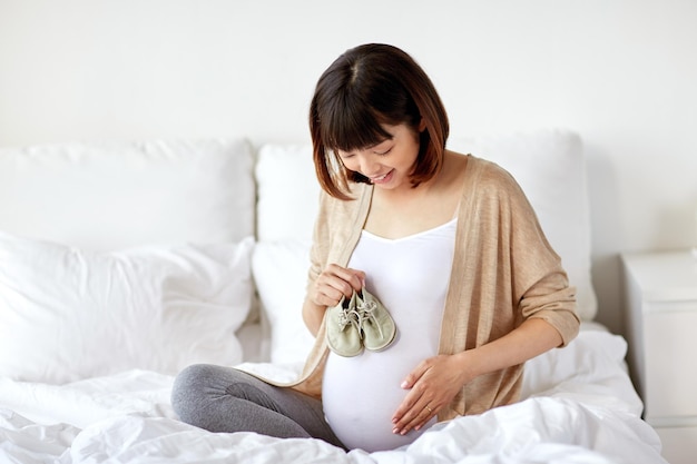 embarazadas personas y concepto de maternidad feliz embarazada mujer asiática con botas de bebé sentado en la cama en el dormitorio de casa
