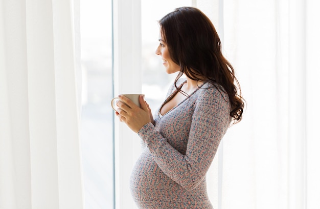 Embarazo, bebidas, descanso, gente y concepto de expectativa: cerca de una mujer embarazada feliz con una taza de té mirando por la ventana en casa