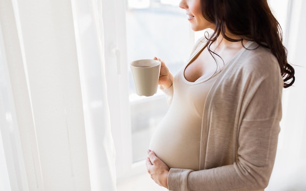 Embarazo, bebidas, descanso, gente y concepto de expectativa: cerca de una mujer embarazada feliz con una taza de té mirando por la ventana en casa