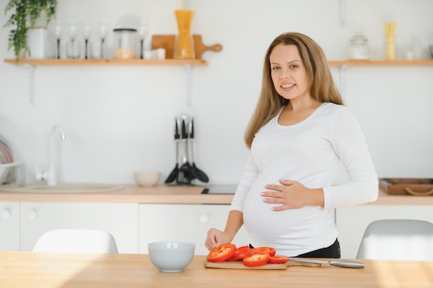 Embarazo, cocinando alimentos, estilo de vida saludable, personas y concepto de expectativa - feliz mujer embarazada con cuchillo cortando pepino y preparando ensalada de verduras en casa